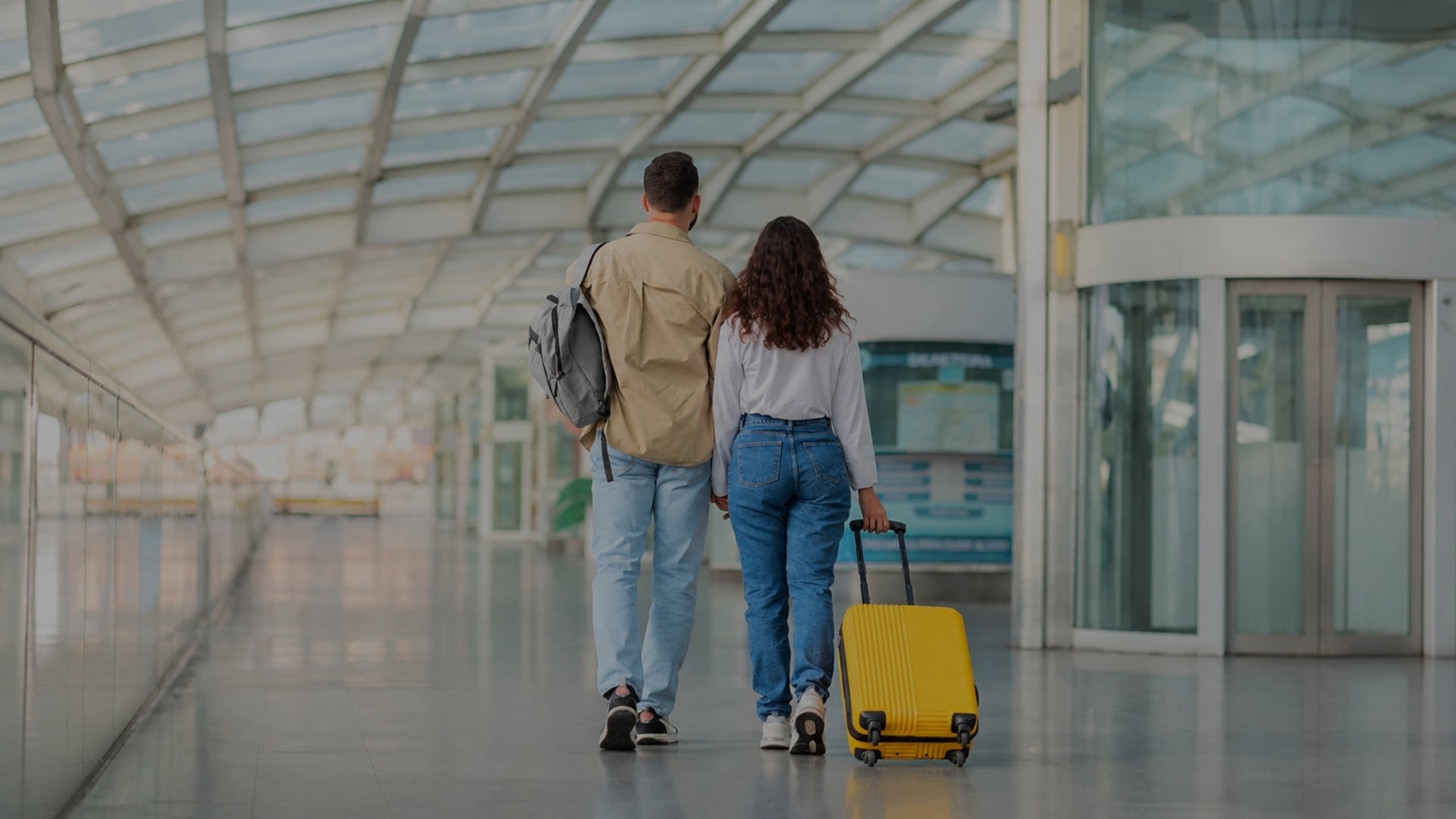 A man and a woman walk away from the camera in an airport terminal, with the woman pulling a bright yellow rolling suitcase. The man wears a beige jacket and light blue jeans with a gray backpack, and the woman wears a white top and blue jeans.