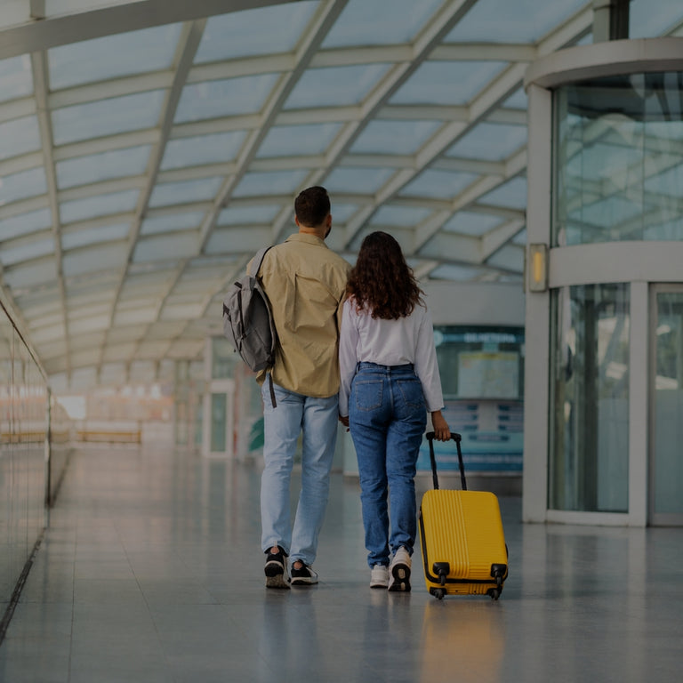 A man and a woman walk away from the camera in an airport terminal, with the woman pulling a bright yellow rolling suitcase. The man wears a beige jacket and light blue jeans with a gray backpack, and the woman wears a white top and blue jeans.
