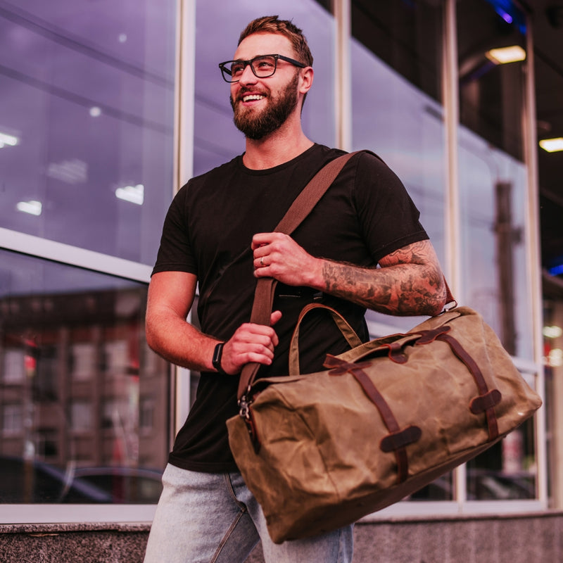 A man with glasses, a beard, and tattoos carries a large brown canvas duffel bag with brown leather straps and trim, standing in front of a modern glass building.