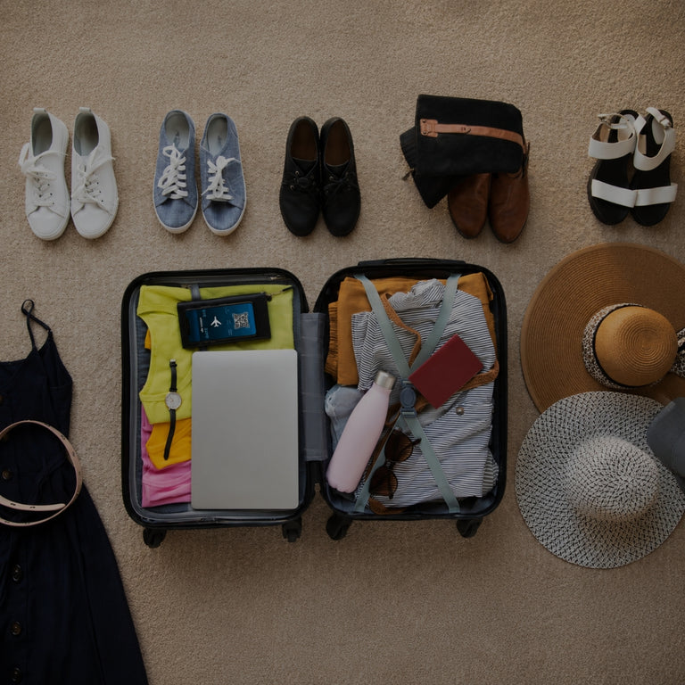 A top-down view of a packed suitcase surrounded by various travel accessories and clothing items on a beige carpet. The suitcase is open and filled with clothes, a laptop, sunglasses, and a water bottle, while surrounding items include shoes, a black top with a belt, a straw hat, a white hat, and a gray beanie.