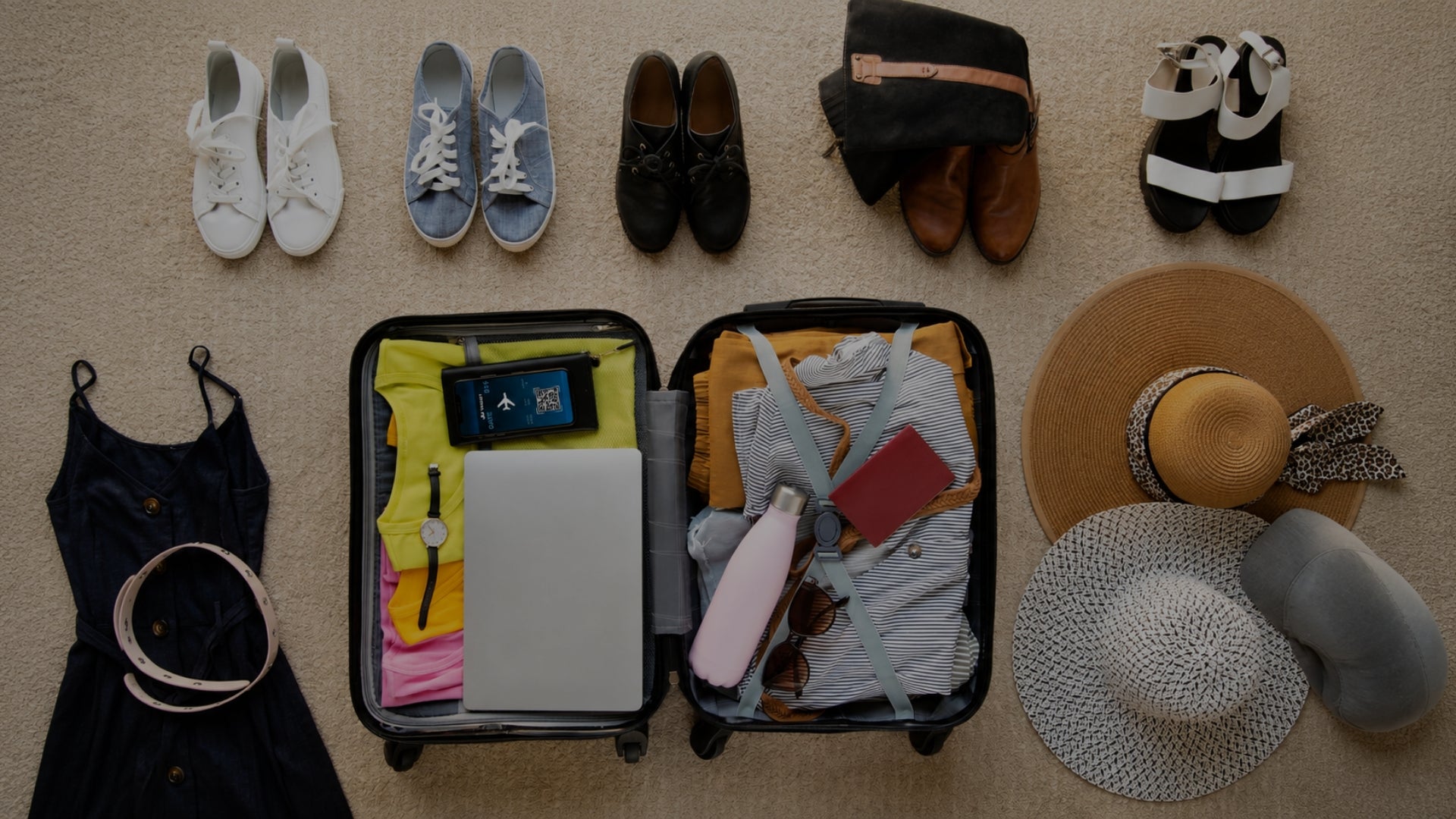 A top-down view of a packed suitcase surrounded by various travel accessories and clothing items on a beige carpet. The suitcase is open and filled with clothes, a laptop, sunglasses, and a water bottle, while surrounding items include shoes, a black top with a belt, a straw hat, a white hat, and a gray beanie.