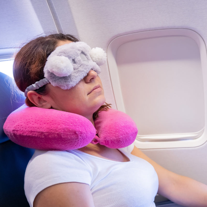 A woman resting on an airplane seat with a bright pink U-shaped travel neck pillow and a gray plush eye mask.